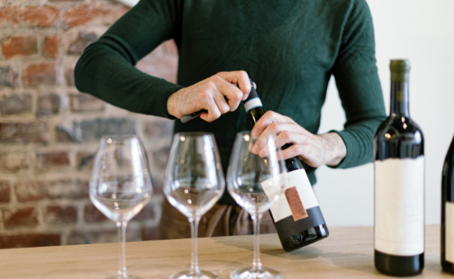 A headless person in a green sweater opening a bottle of red wine with a waiter's corkscrew